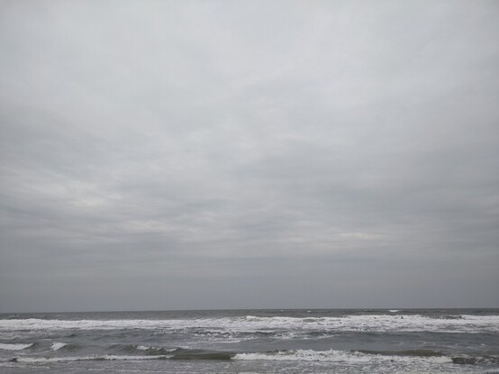 Photo: Breaking waves and an expanse of ocean reach to the horizon. The ocean is gray and flecked with white foam where the waves are breaking. The sky is covered by gray clouds that are very nearly uniform in color. About seven-eighths of the photo are gray sky, while the ocean makes up only one-eighth of the scene. 