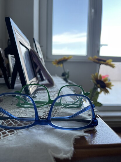A pair of blue glasses in front of a pair of green glasses, set on a table with picture frames, faux sunflowers, windows, sky in the background.